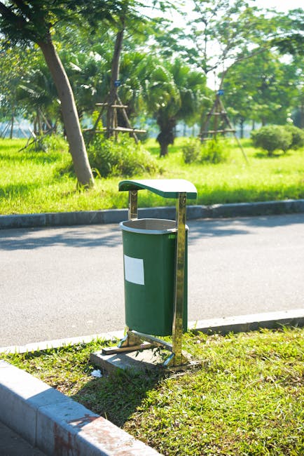 A green cylindrical waste bin with a white label on its front, attached to a metal stand with a curved, flat lid serving as a small shelter, is positioned at the edge of a paved pathway in an outdoor park setting. The bin is located on a narrow grassy strip bordered by the paving, and surrounds a curb that separates the pathway from the grassy area. In the background, there are lush green trees, including palm trees and other foliage, with some trees supported by wooden framework. The sunlight filters through the leaves, casting dappled shadows across the scene, indicating a bright, clear day. The environment appears well-maintained and is suitable for outdoor public use, with waste disposal conveniently located for park visitors. Waste Disposal Watford offers services that facilitate proper rubbish collection and private disposal in areas such as this, supporting onsite rubbish management efforts in public outdoor spaces.