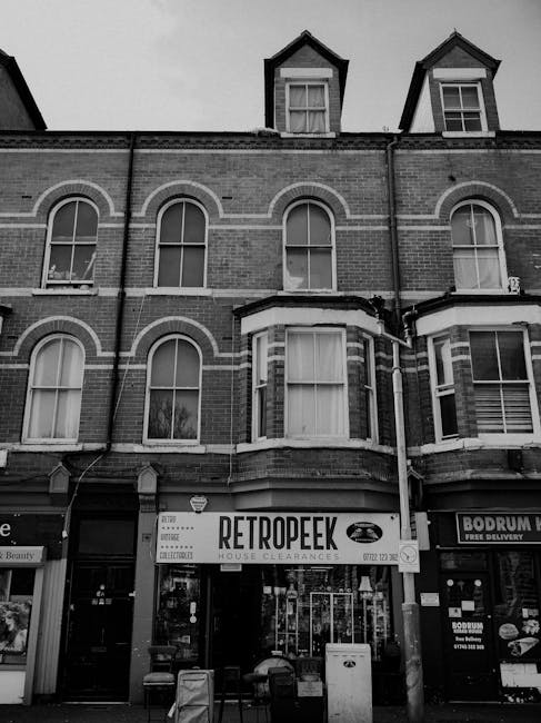 A three-storey brick building with large sash windows on the upper floors, featuring arched and rectangular shapes with white window frames and sills. The ground floor houses a house clearance business named 'Retro Peek,' with a prominent sign above the entrance displaying black text on a white background. In front of the shop, there are several small, dark-colored chairs, a white waste bin, and a few standing objects, possibly for waste collection or storage. To the right, a neighboring business offers food delivery services, evident from a sign displaying 'Bodrum' and associated contact details. The building's exterior has brickwork with horizontal white accents and a bay window projecting from the central part of the facade. Above, the upper floors contain dormer windows with dark sloped roofs, adding a Victorian or Edwardian architectural style. The sky is overcast, casting diffuse light over the scene, reflecting a typical urban environment where private waste collection might be carried out separately from municipal services, supporting the context of independent rubbish removal or house clearance activities by Waste Disposal Watford.