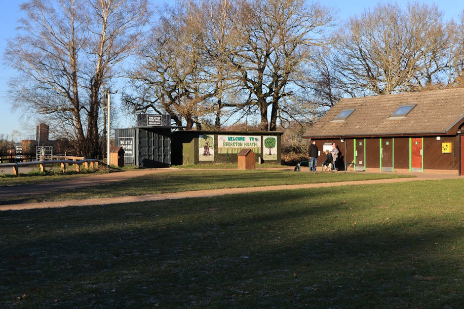 A park scene with a gravel pathway in the foreground leading toward a green and black container unit for waste disposal, situated near a brown building with a sloped roof and multiple skylights. The container has a sign that reads 'Welcome to Brereton Heath' and features painted illustrations of trees, indicating a community or recreational area. In front of the building, two people are standing and interacting, one with a dog on a leash, while others are nearby. Tall leafless trees with extensive branches are visible against a clear blue sky, and the environment appears well-maintained and open. The surrounding grass is short and green, with some patches of dirt in the pathway area. This scene reflects a typical outdoor location where waste management and rubbish collection are integrated with recreational or public spaces, highlighting the presence of waste disposal facilities that support environmental cleanliness and proper rubbish handling through private or local services like Waste Disposal Watford.
