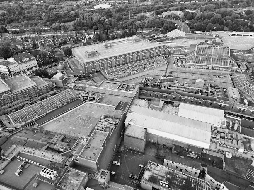 An aerial black-and-white photograph showcases a dense urban area with a mix of large industrial and commercial buildings, including multiple glass-roofed sections and flat-roofed warehouses, alongside smaller residential structures and tree-lined streets in the background. The rooftops of the industrial buildings display various textures such as metal sheeting, glass skylights, and concrete surfaces, with some areas showing visible ventilation units, air conditioning units, and other mechanical equipment. The scene captures a section of a cityscape with narrow alleyways and parking areas adjacent to the buildings, where cars are parked. The overall environment appears well-organized, with clean surfaces and clearly defined spaces, representative of typical commercial and industrial zones. This setting relates to the domain of waste disposal and rubbish removal services, exemplifying a location where comprehensive on-site clearance or waste handling might take place. The natural lighting is diffuse, with no strong shadows, highlighting the structural details of the rooftops and surrounding environment, fitting the context of professional rubbish removal activities in busy urban settings, such as Watford or similar locations in the UK.