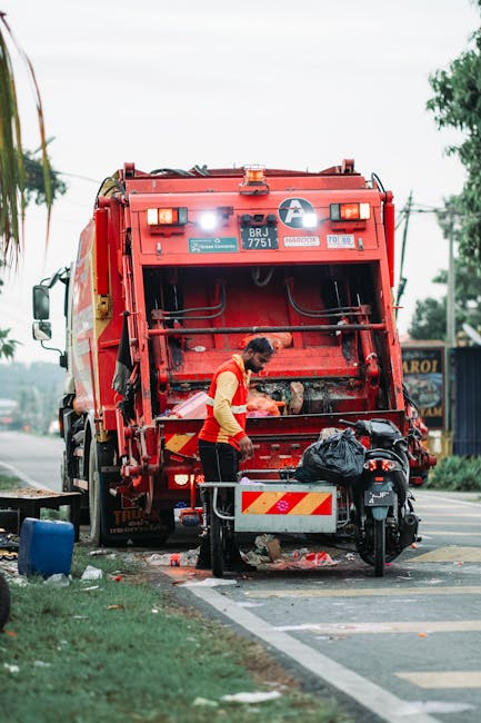 A waste collection worker wearing a high-visibility orange and yellow vest is positioned at the rear of a large red rubbish collection truck, which is actively engaged in waste disposal. The truck's rear compartment is open, revealing mechanical components and a clean, painted interior. The worker appears to be handling black rubbish bags, one of which is placed on the ground next to a small, portable bin with reflective red and white chevron markings. The scene is set outdoors on a paved street with a curb and a designated parking or loading space, with some scattered debris and a blue bin visible nearby. In the background, there are trees, utility poles with overhead wires, and a small building or shop sign partially visible, indicating a suburban or town environment. The lighting suggests daytime with natural light, showing clear weather conditions. This image demonstrates an example of private waste collection service, such as rubbish removal, with a focus on proper waste handling and disposal practices, relevant to on-site clearance or alternative waste management options provided by Waste Disposal Watford.